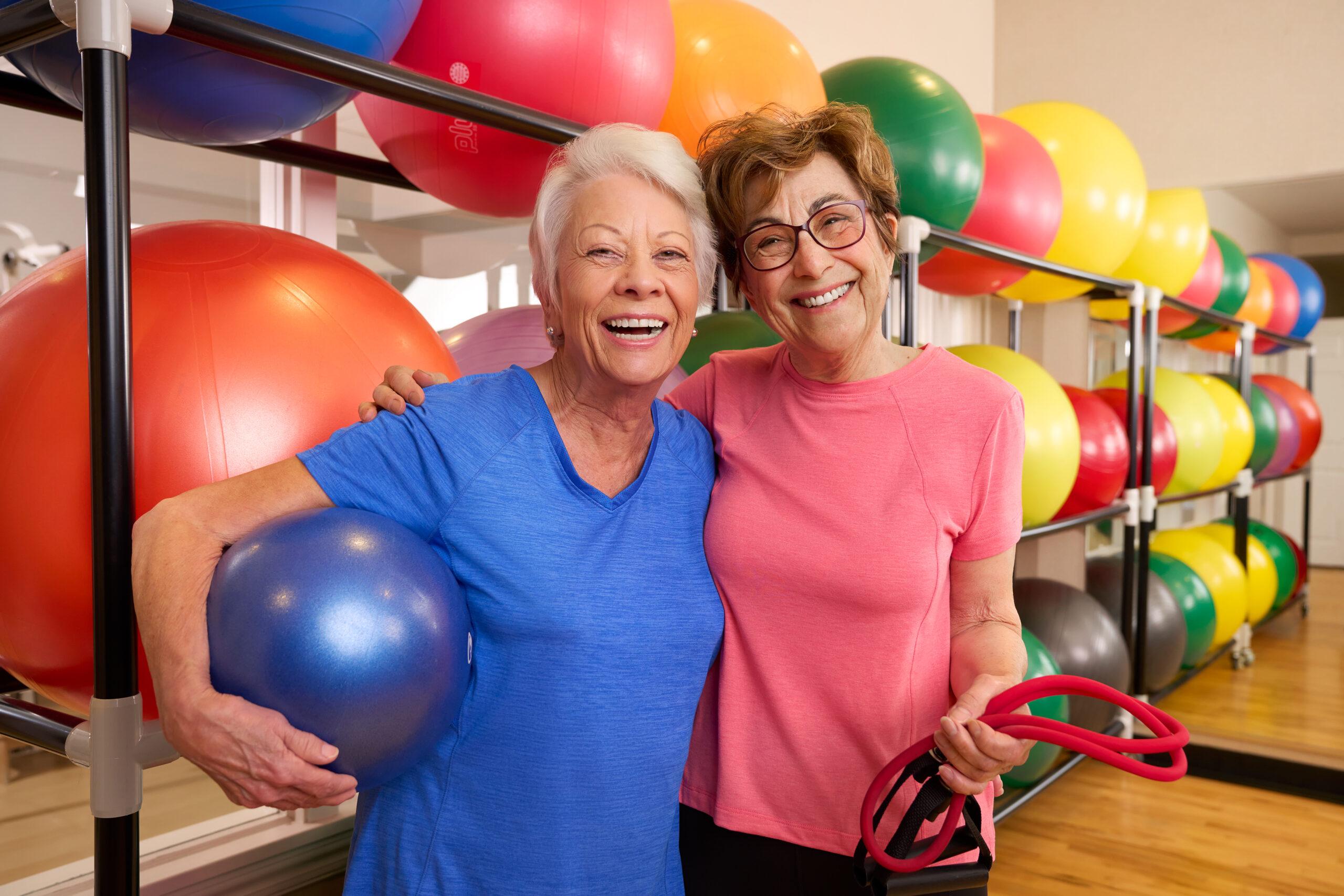 Two older women smile in front of a group of colorful exercise balls