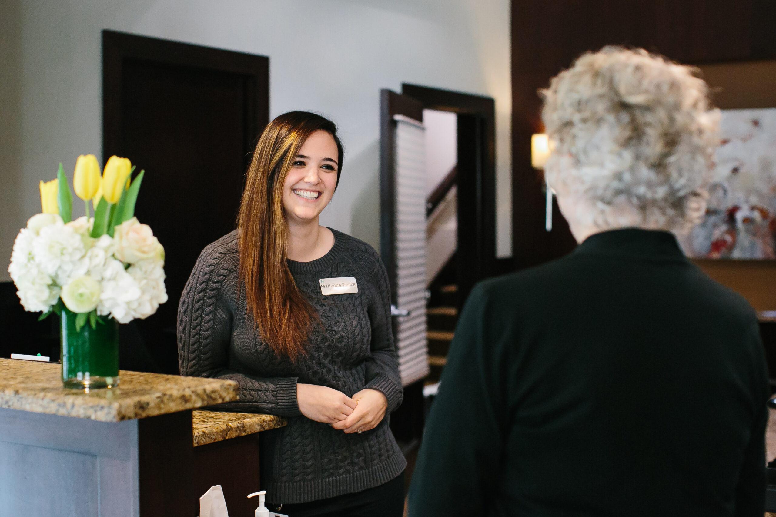 A front desk worker smiles while talking to an older person, who is faced away from the camera