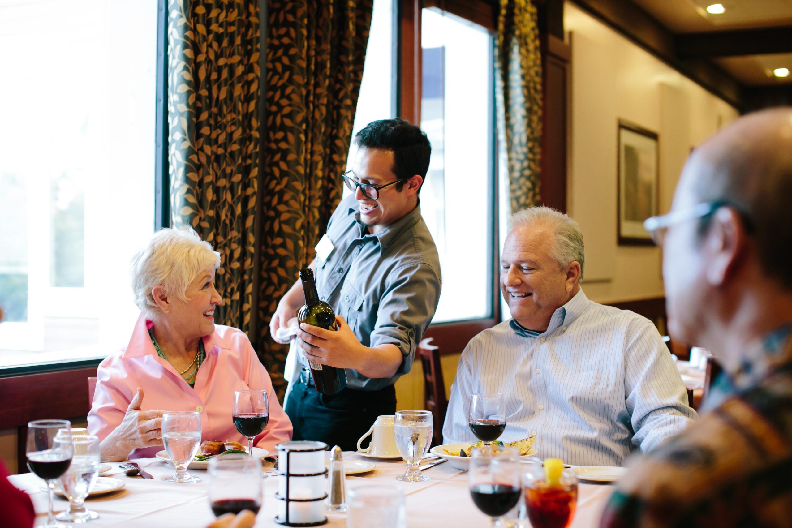 A playful waiter pours a glass of wine for an older woman sitting at the table with friends