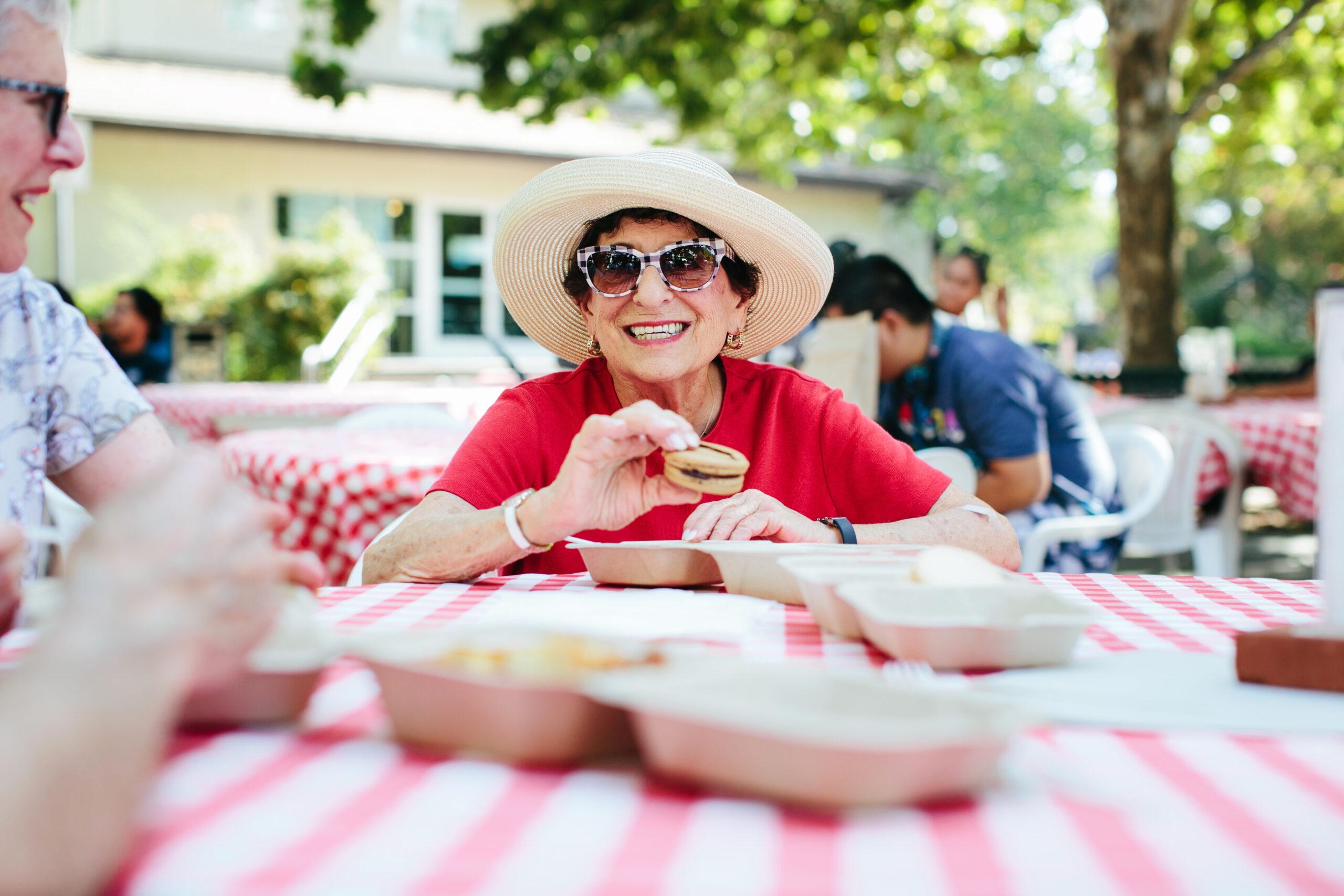 An older woman wearing a hat and sunglasses enjoys a snack at a picnic table