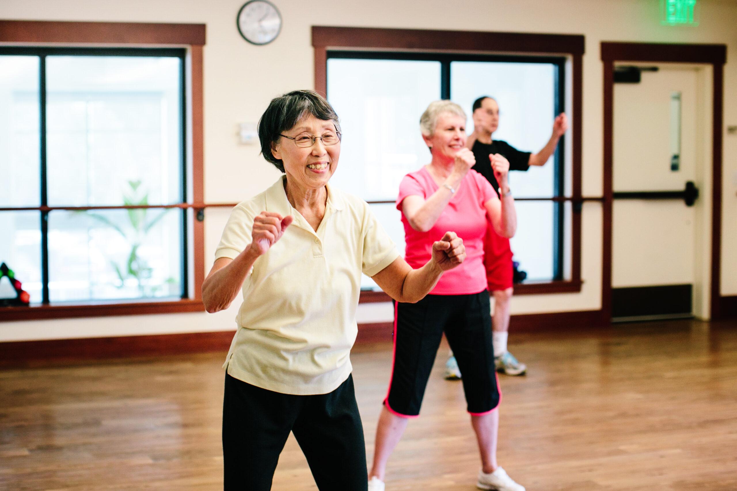 Three seniors move around at a group fitness class. The camera is focused on a smiling woman in the foreground