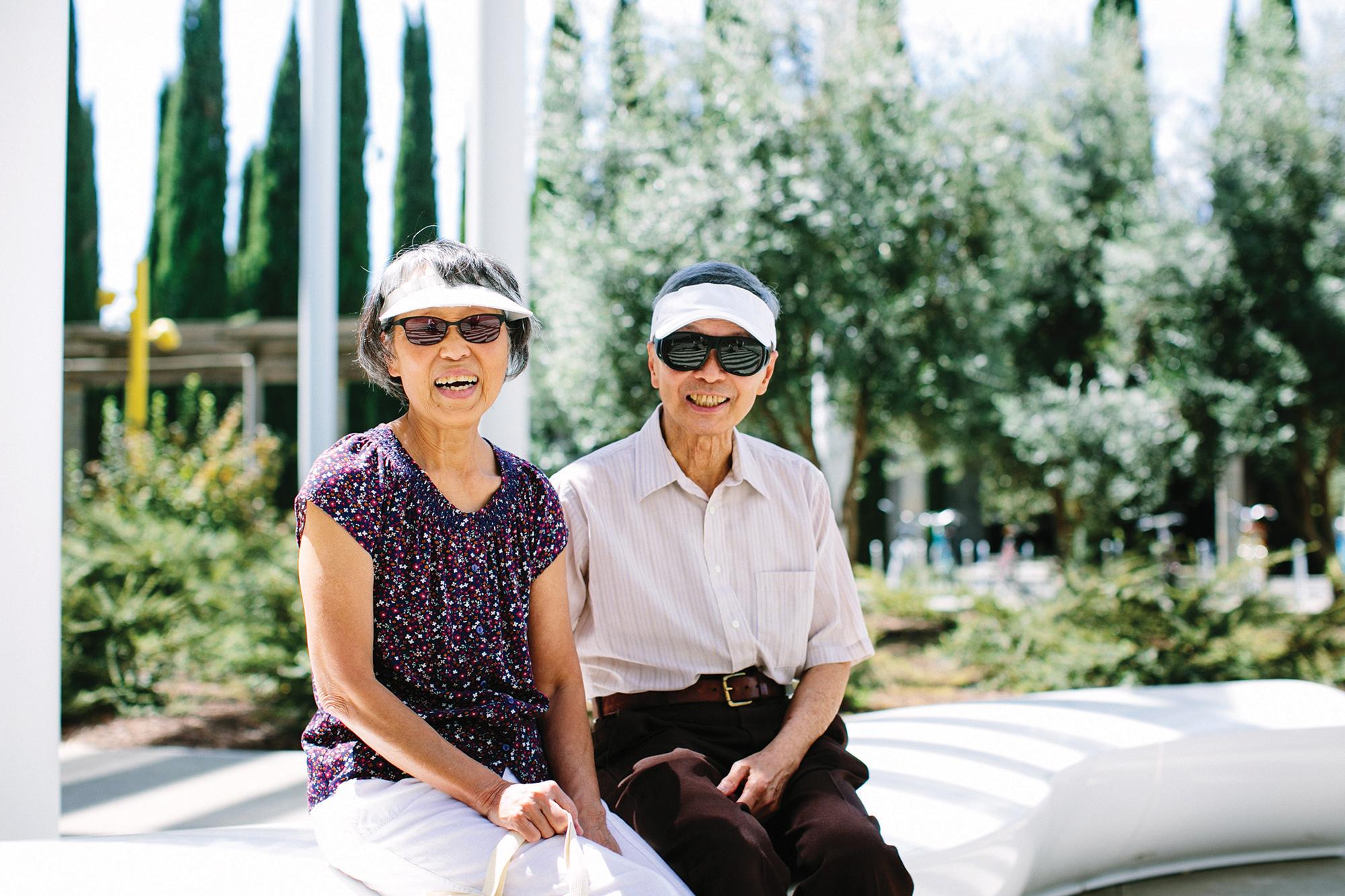 A smiling older couple wearing sunglasses and golf visors sits outdoors.